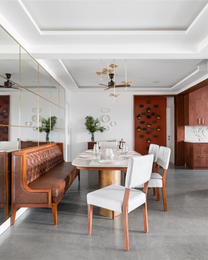 The is anchored by a live-edge wooden table paired with upholstered chairs and a wooden bench. A brass-accented chandelier casts a soft glow while a mirrored wall reflects light from the French windows. Natural stone flooring and details like linen runners and ceramic tableware enhance the room’s luxury; Photography by Justin Sebastian