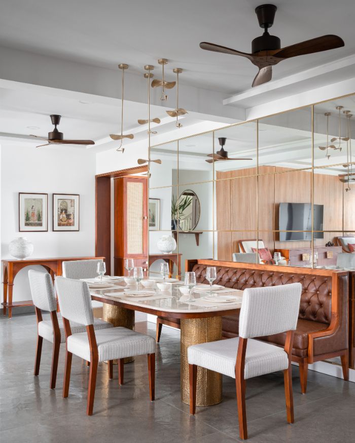 The dining area is anchored by a live-edge wooden table paired with upholstered chairs and a wooden bench. A brass-accented chandelier casts a soft glow while a mirrored wall reflects light from the French windows. Natural stone flooring and details like linen runners and ceramic tableware enhance the room’s luxury; Photography by Justin Sebastian