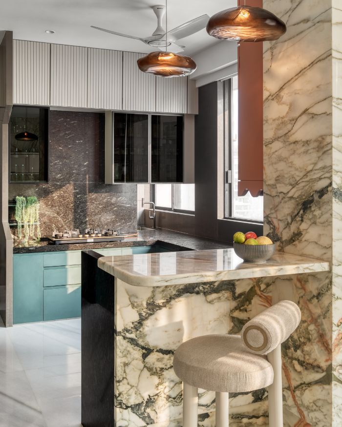 The kitchen unfolds in artful layers as veined marble from The Quarry wraps the breakfast counter and bar in fluid movement. Granite by The Classic Marble Company and lighting from VLights complete the space with timeless elegance;  Photography by Pulkit Sehgal