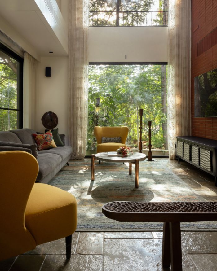 A tall, airy living room is anchored by a grey sofa and mustard-yellow armchairs around a round coffee table. Floor-to-ceiling windows flood the space with natural light and offer a lush, uninterrupted view of the garden just beyond; Styling by Fymin Naif and Nimitha Harith; Photography by Justin Sebastian