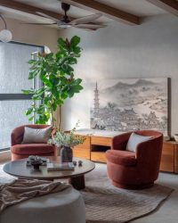 In the living room, sunlight filters through a ten-foot barringtonia from Ugaoo, its leaves casting shifting shadows on a vintage Japanese art reprint. The space is layered with soft furnishings from Altrove, a Rugberry rug underfoot, and sculptural furniture by EK Design. Featuring chairs from West Elm and a floor lamp from Orange Tree, coffee table books from Bookchor, vases from Altrove and ceiling light from Orra. The walls, finished in Lepan limewash and framed by wood beam ceilings; Photography by Kuber